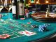 A sophisticated casino table setup featuring various wine bottles and glasses alongside poker chips, cards, and a roulette wheel, illustrating the luxurious pairing of wines with casino games.